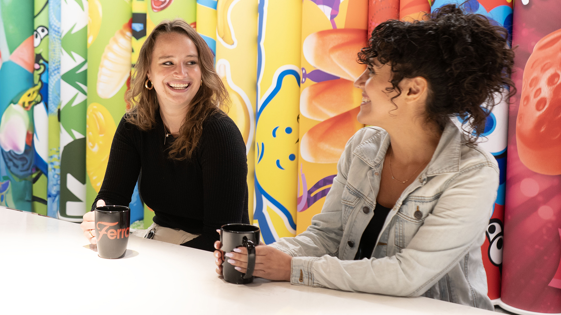 Two female employees drinking coffee and talking in front of a colorful Ferrara branded background
