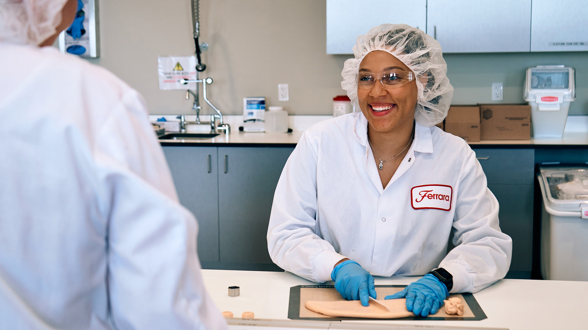 Smiling female employee scientists wearing a hairnet white coverall with her name Jaylene and blue gloves while working with candy in a lab