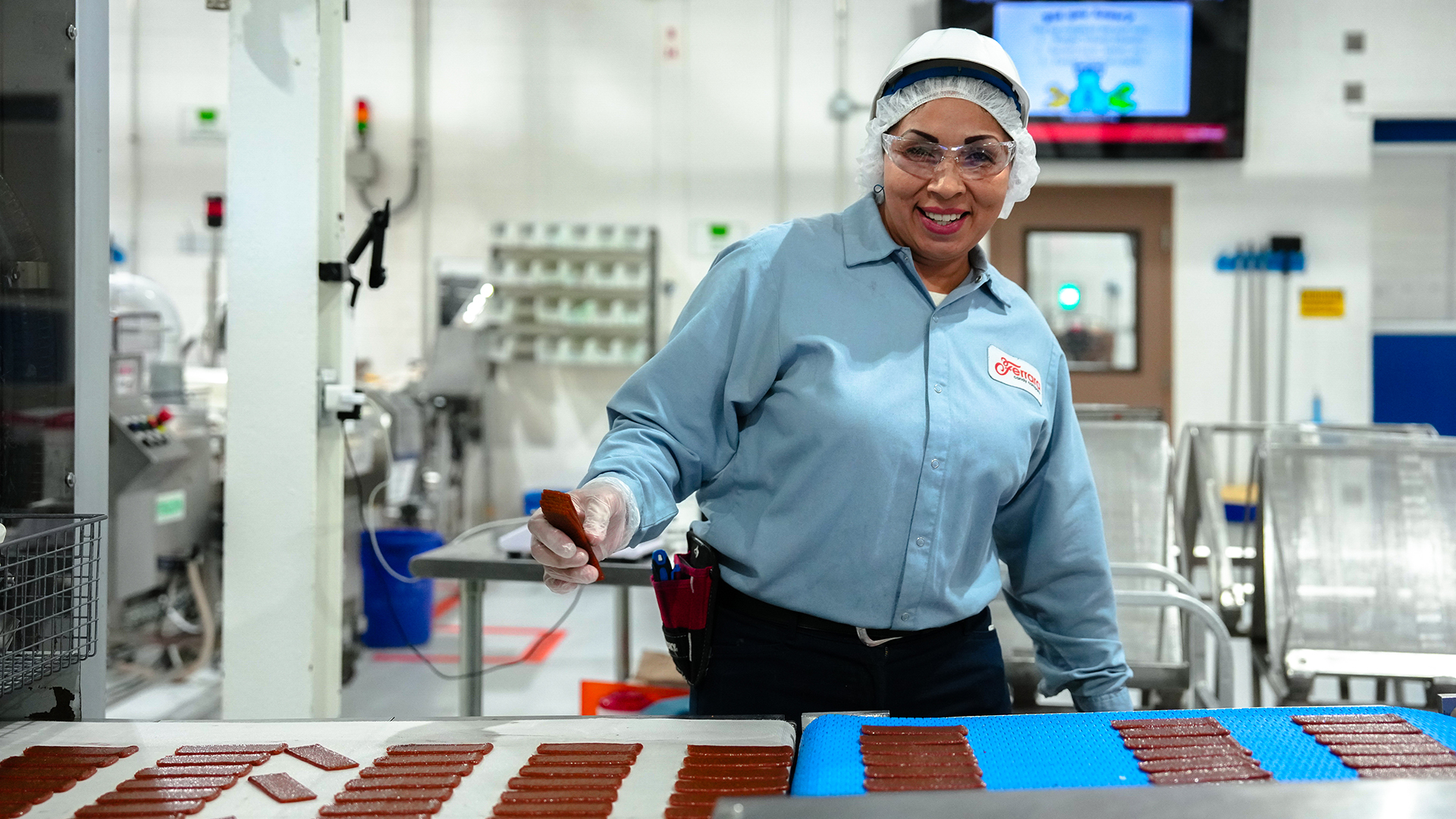 Female Ferrara employee wearing a hairnet, hard hat, latex gloves, and logoed blue shirt holding a fruit bar and smiling standing behind a line of fruit bars