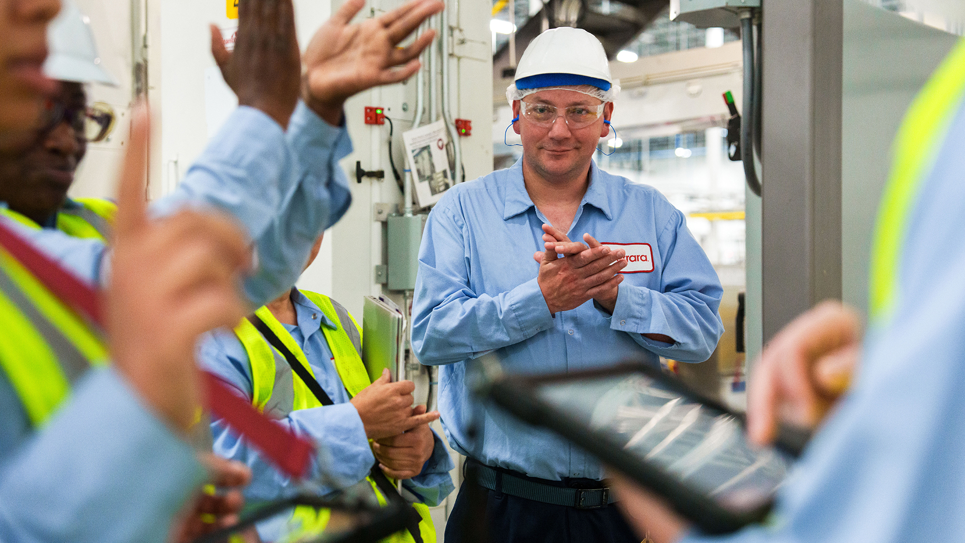 Focus on male employee wearing hair net, hardhat, and blue logoed shirt with the name Todd clapping, while other employee hands clap in foreground