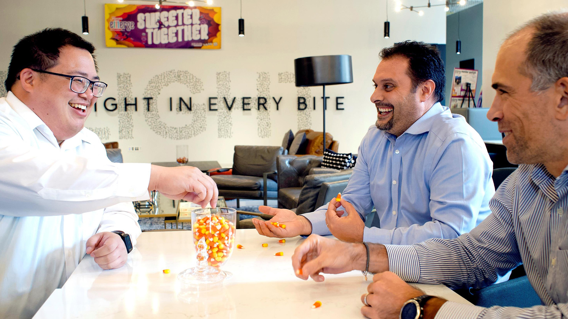 Three male employees in a Ferrara breakroom sitting at a table smiling and eating candy corn. Sweeter Together logo on wall in background with the words ...ight in every bite.