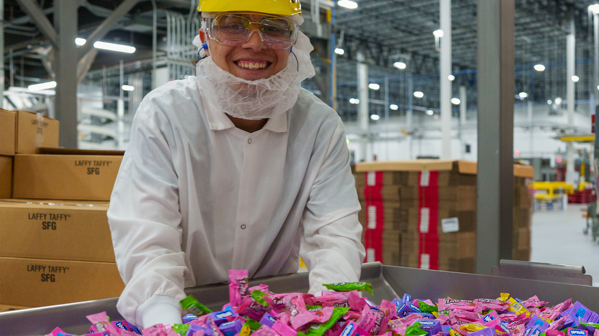 Male employee wearing a hard hat, beard net and white over coat with his hand in a large bin of Now and Laters individually wrapped candies
