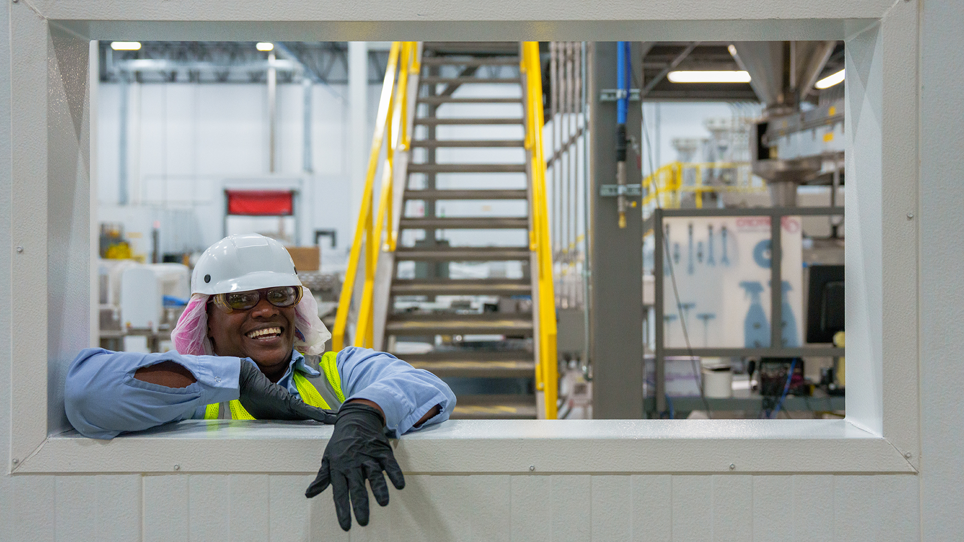 Smiling female employee wearing hairnet, hard hat, gloves and hi viz vest leaning through window opening in plant and smiling