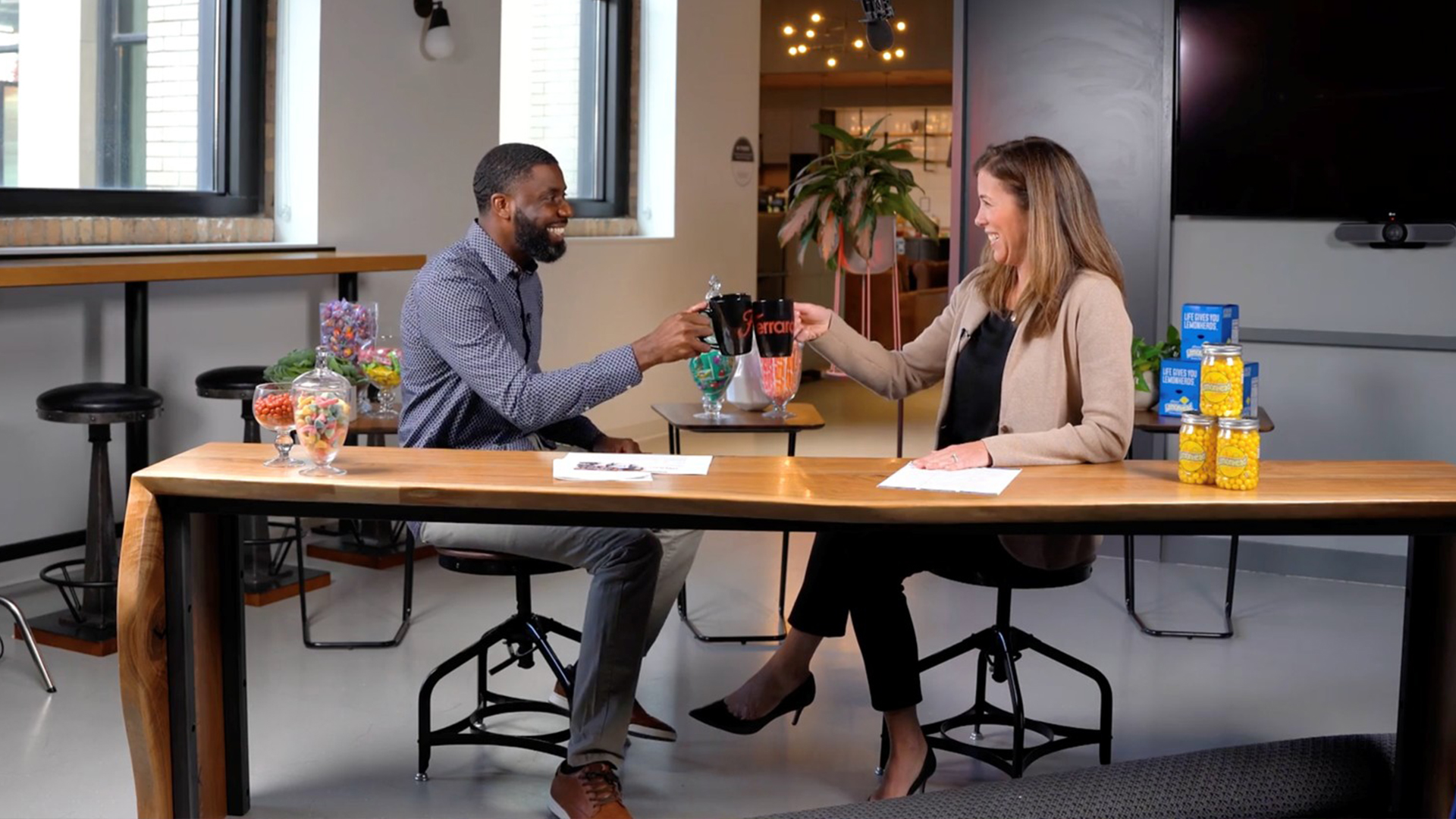 Male and female clinking Ferrara branded coffee mugs while sitting at a table getting ready of a video taping. Microphone can be seen hanging down at top of image.