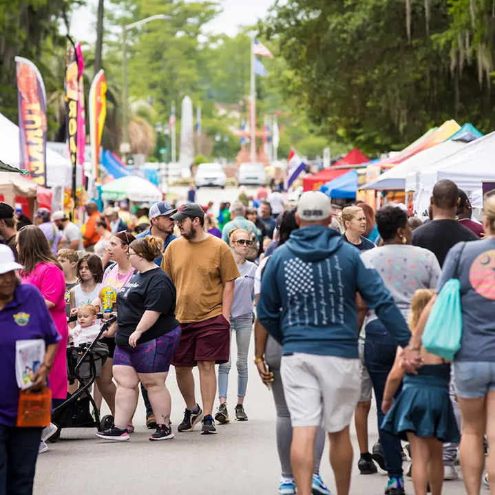 a large crowd of people walking through a festival with vendor tents and flags on either side of the street